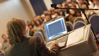 Eine Frau steht vor einem Publikum an einem Rednerpult mit Laptop.